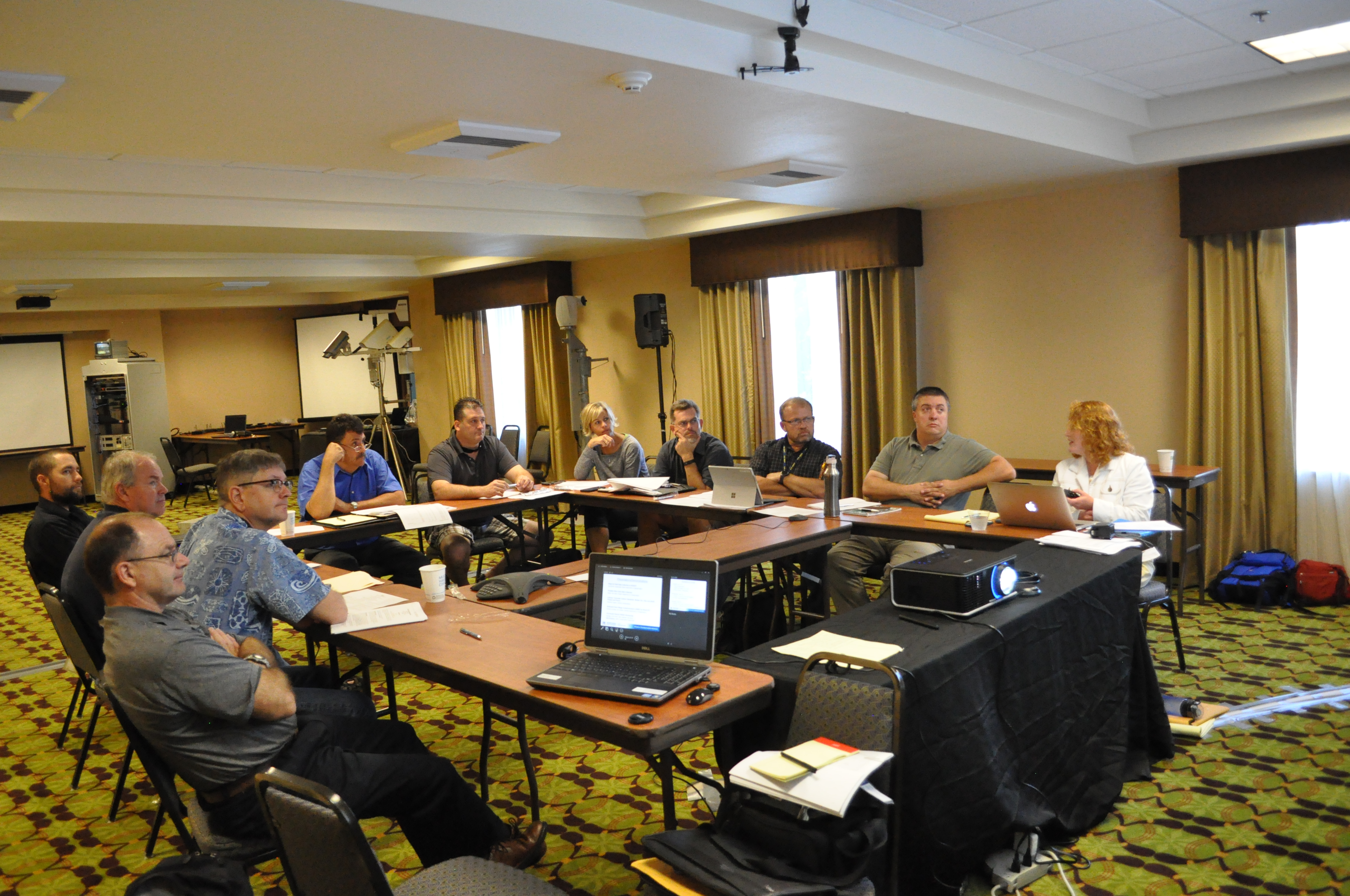 A group of people seated at rectangular tables arranged in a U shape, projector.