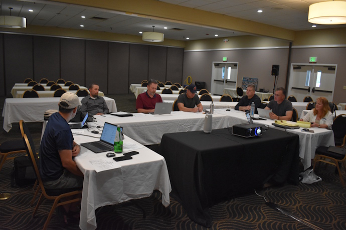 Seven people at a meeting sitting at tables in a U shape configuration with a projector on a table in the middle. One person is speaking while the others are listening to him.