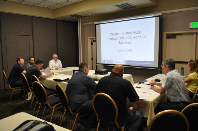 A group of people seated at tables in a U shape with a projector screen in front, slide on screen with text: Western States Rural Transportation Consortium Meeting.