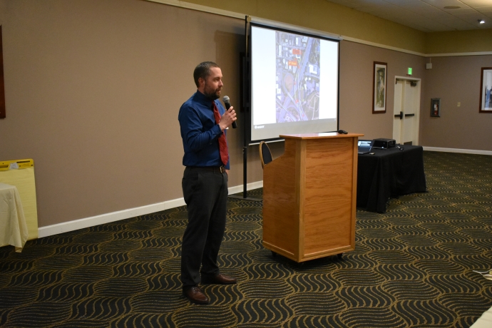 A man holds a microphone and speaks to an audience in front of a projector.