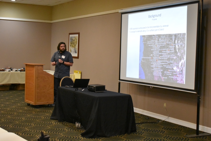 A man holds a microphone while standing beside a projector screen.