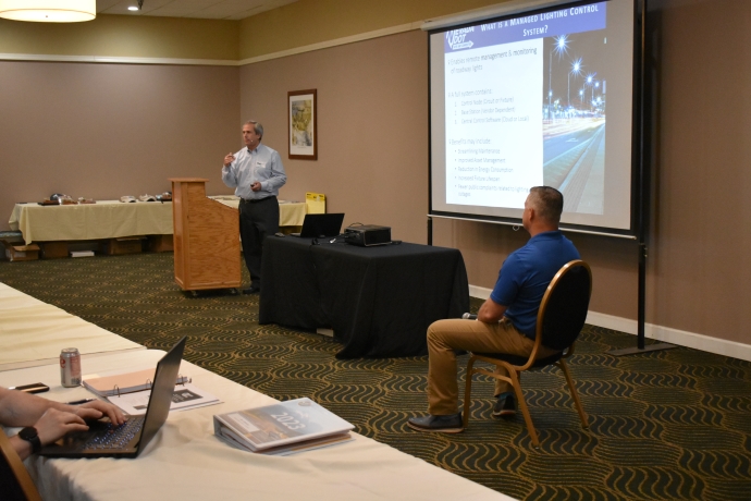 A man presents to the left of a projector screen. Another man is seated across from him.