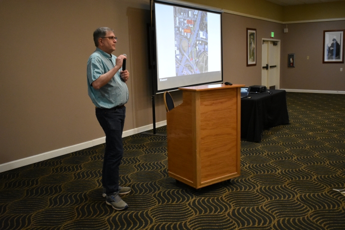 A man holds a microphone and speaks to an audience in front of a projector.