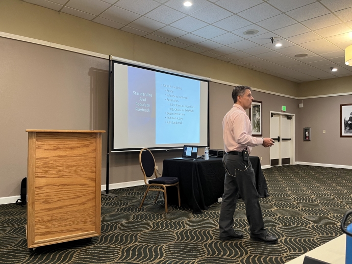 A man walks in front of an audience while presenting material displayed on a projector screen.