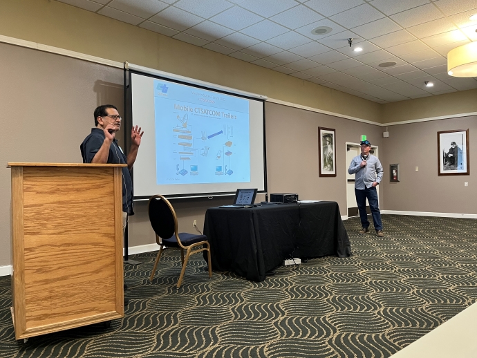 Two men stand on either side of a projector screen. The man on the left behind a podium is presenting to an audience.