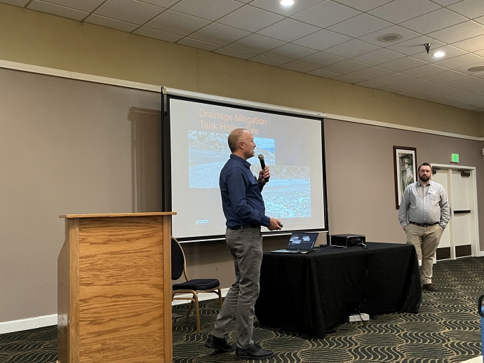 Two men stand on either side of a projector screen. The one on the left is speaking into a microphone.