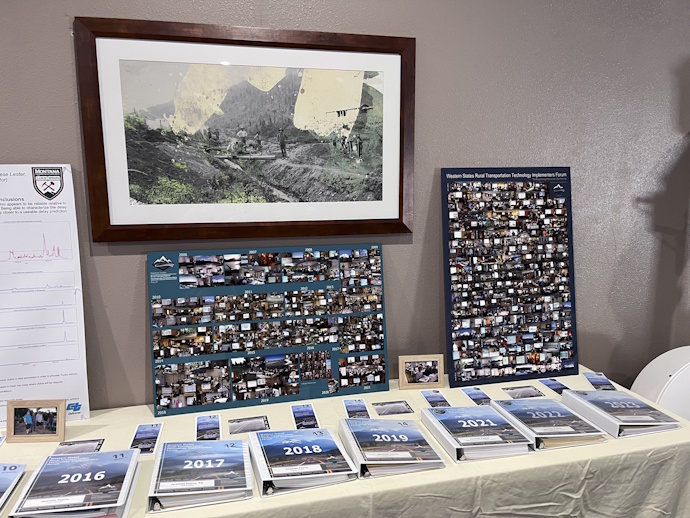 On a table: two large photo collages, Forum three ring binders from 2016 to 2023, brochures and postcards.