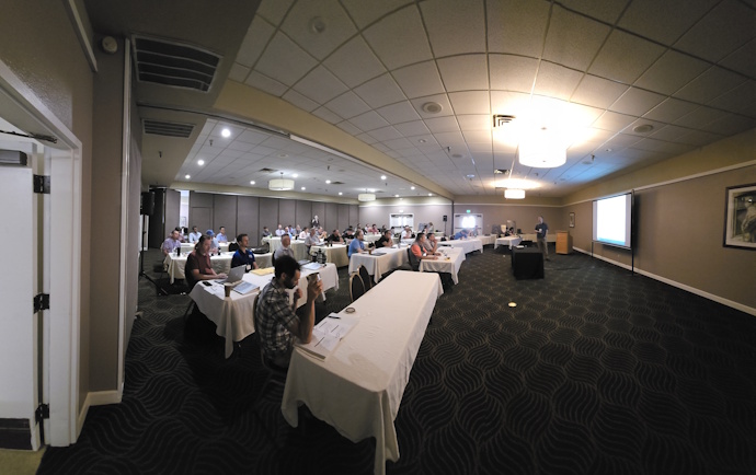 Front view of an audience seated classroom style listening to a speaker standing in the front of a medium sized room.