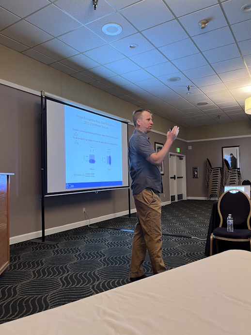A man gestures with his right hand while speaking beside a projector screen.