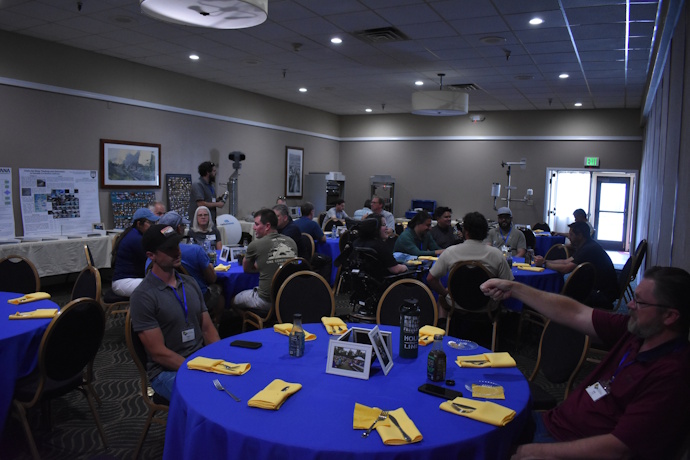 Groups of people seated at round tables covered by bright blue tablecloths with yellow napkins. A man in the front right has his arm raised while talking to a man across the table from him. Photo collages, a poster, and equipment are seen in the background along the wall.