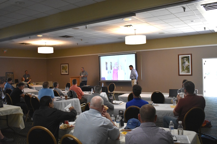 An audience looks to the front of a room where three men are standing and facing them. In the middle, two women are shown on a screen. The one on the left is wearing a pink dress and pink baseball cap. The one on the right has a yellow cap.