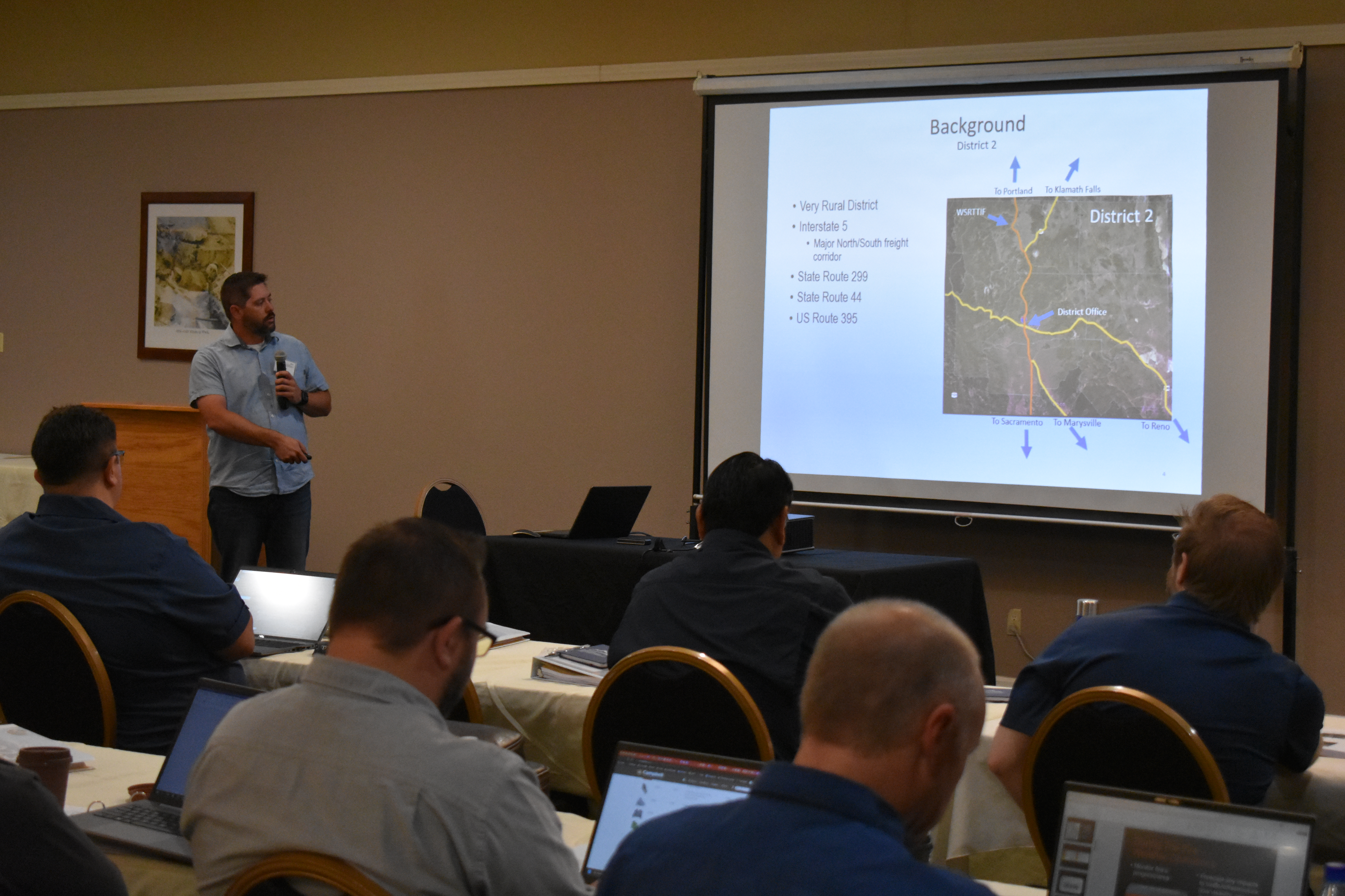 A man holds a microphone and speaks to an audience in front of a projector.